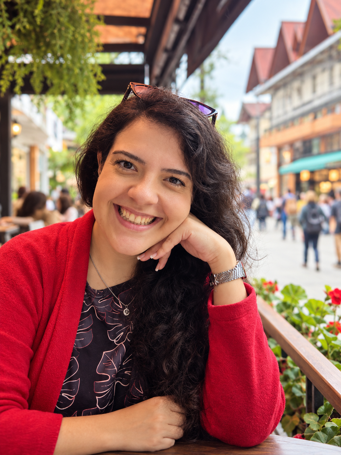 Bibi sorrindo, vestida de vermelho, com o queixo apoiado na mão, em uma rua com lojas ao fundo